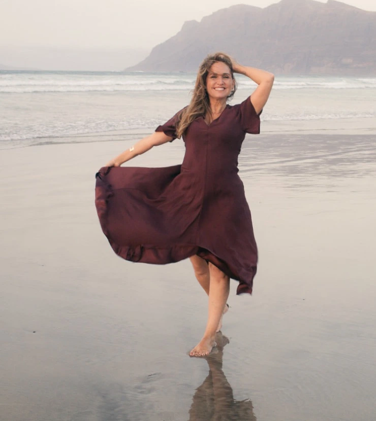 Fayna Curbelo sonriendo y moviendo su vestido en la orilla de Famara, con el mar y los acantilados al fondo.