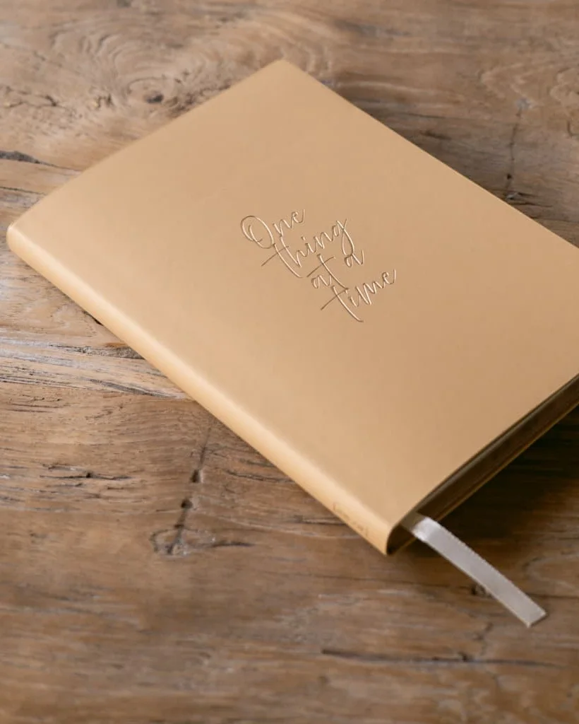 Libreta color arena con la frase «One thing at a time» sobre una mesa de madera.