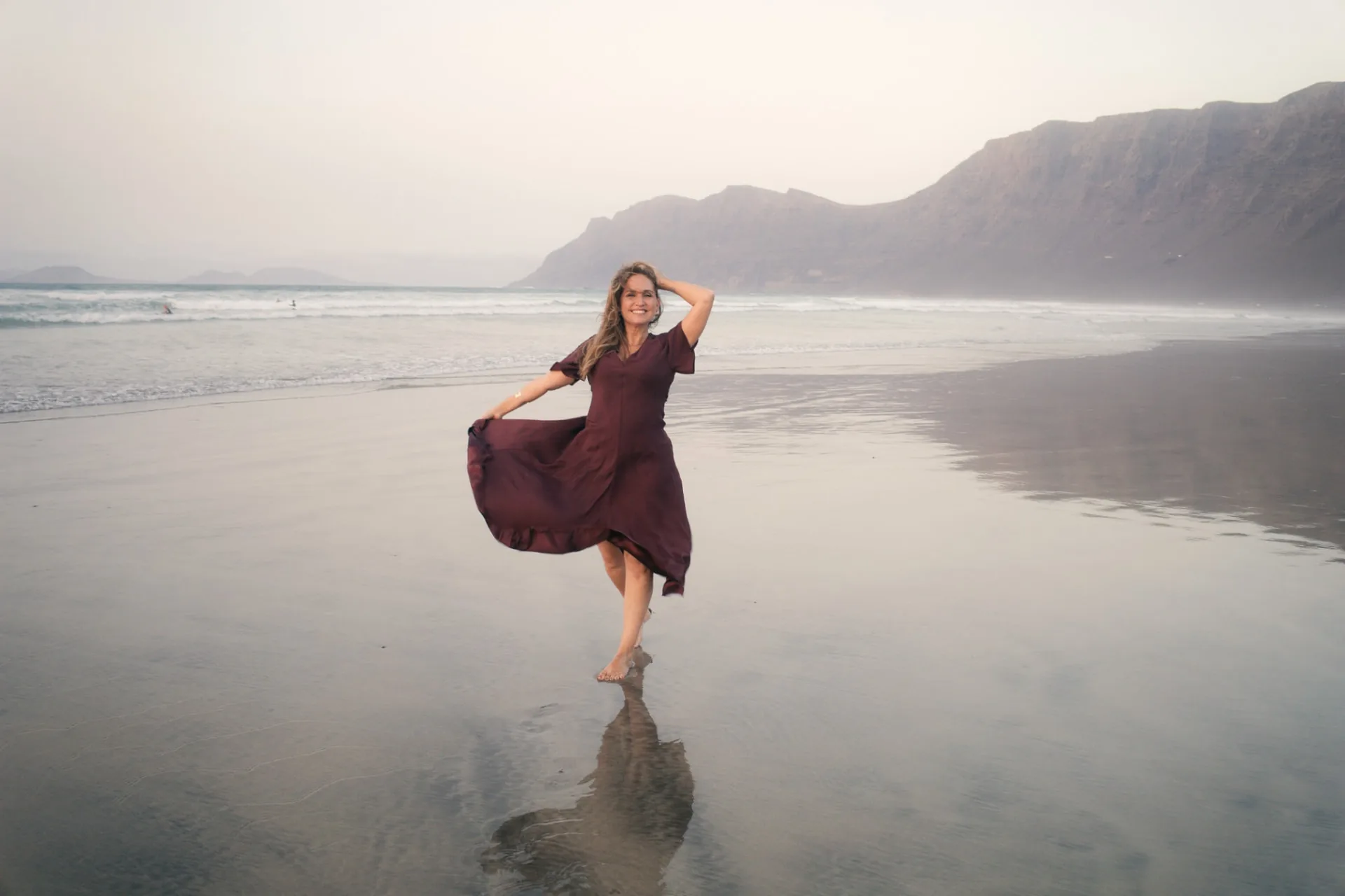 Fayna Curbelo descalza en la orilla de Famara con un vestido color vino, sosteniendo el vuelo de la falda, con su reflejo en la arena mojada y los acantilados al fondo.