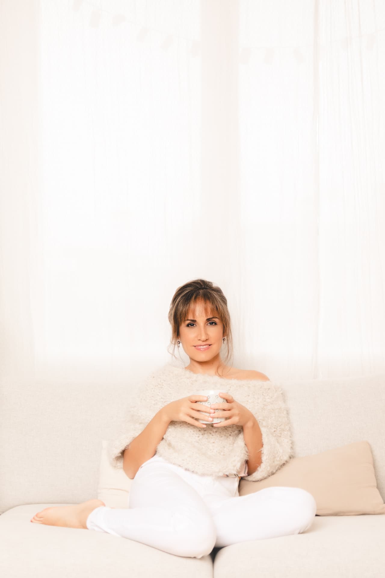 Fayna Curbelo sentada con las piernas cruzadas en un sofá claro, con una taza entre las manos.